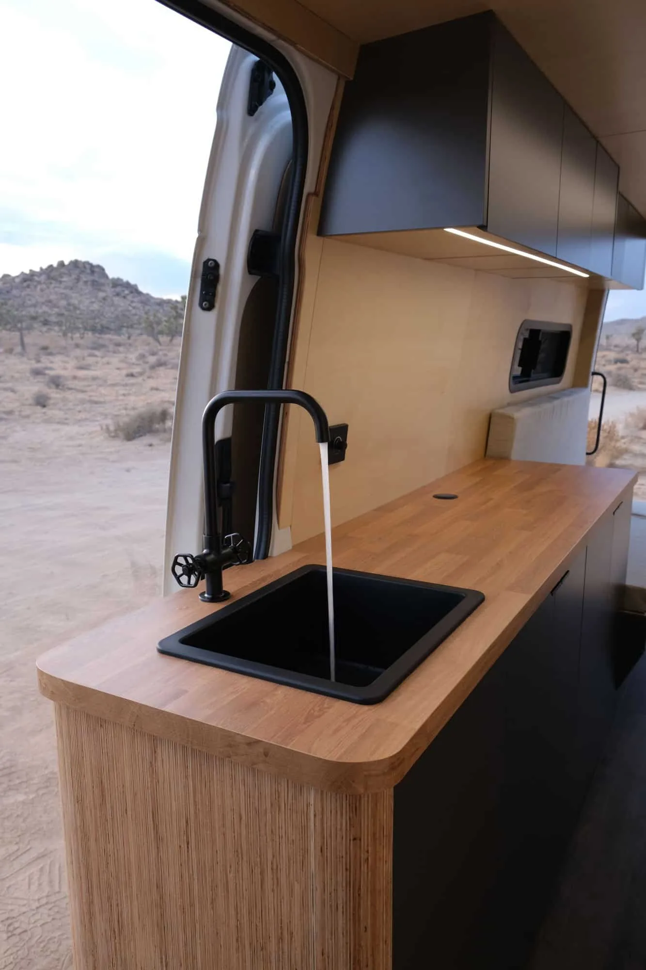Galley inside the converted van — oak worktop, black sink and tap, birch ply cabinets, desert through the open side door